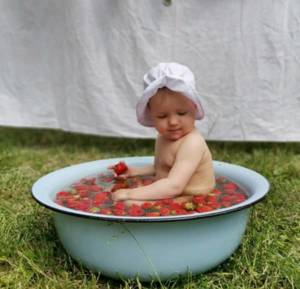 bébé dans une bassine d'eau avec des fraises, dans l'herbe devant un drap blanc